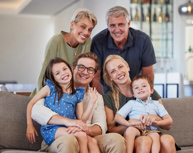 Three generations of a smiling family sitting together on a living room sofa, highlighting family connection, comfort, and healthy smiles.