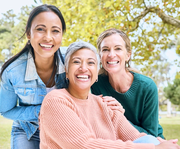 Three smiling women posing together outdoors in a park, representing friendship, wellness, and healthy smiles.